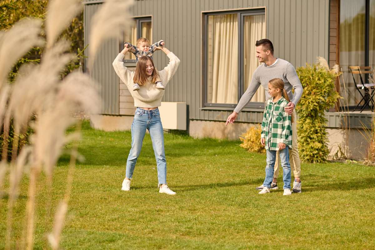 Games. Young joyful long-haired woman in casual clothes holding baby on neck taking step and man hugging daughter pointing finger at lawn playing spending leisure time outdoors